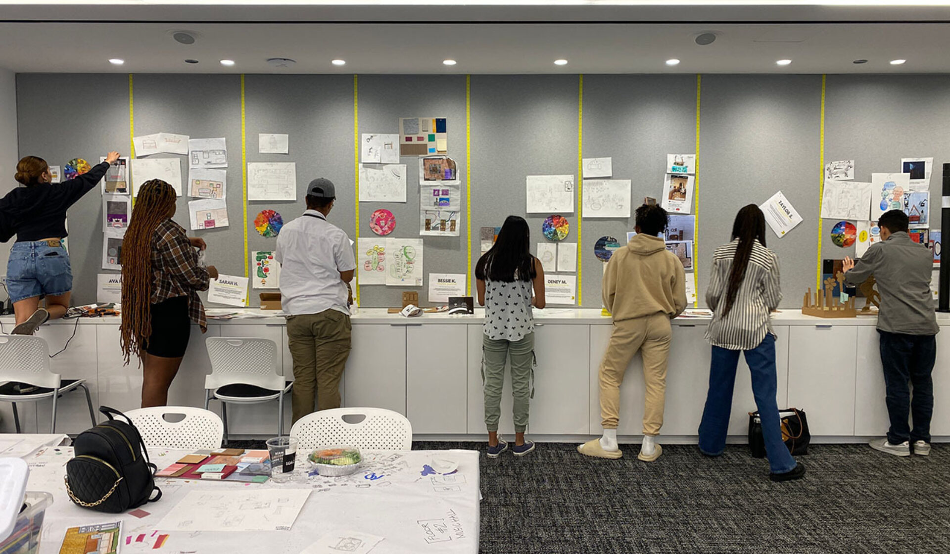 A group of diverse individuals interact with art presentations on a wall and table in a creative workspace, focusing on their work.