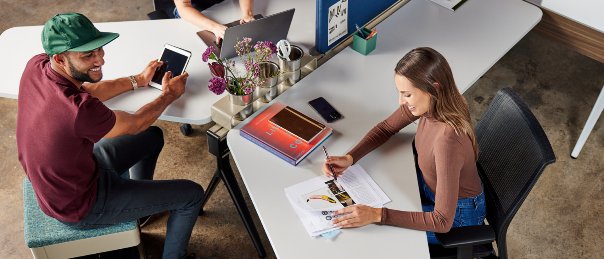 A man sits on a stool using a tablet, while a woman writes at a desk with printed materials and a laptop, in a modern workspace.