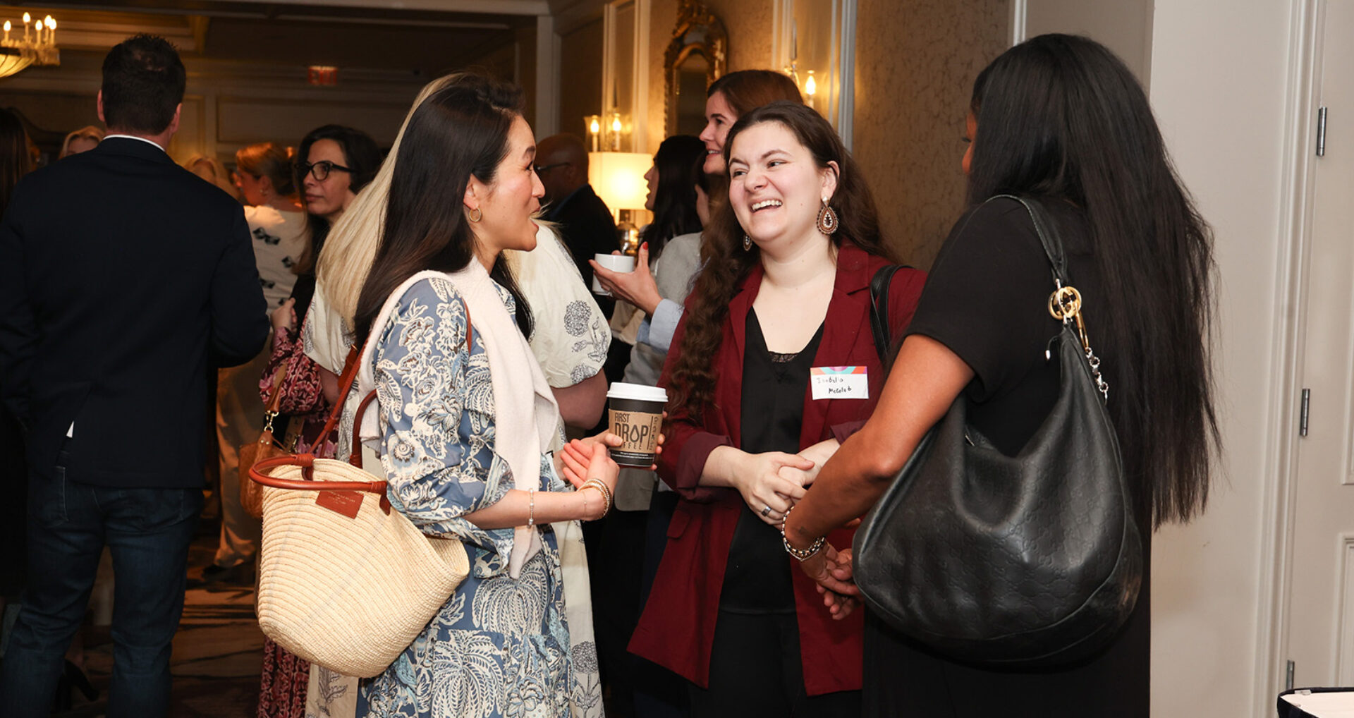 Three women laugh and chat at a crowded networking reception; one holds a coffee cup, another wears a woven tote, warm chandelier light.