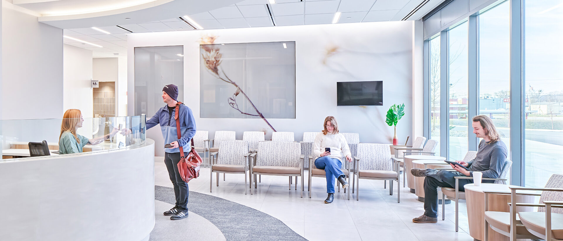 Bright modern medical waiting room with curved reception desk, a man checking in, two people seated using devices, floor-to-ceiling windows.