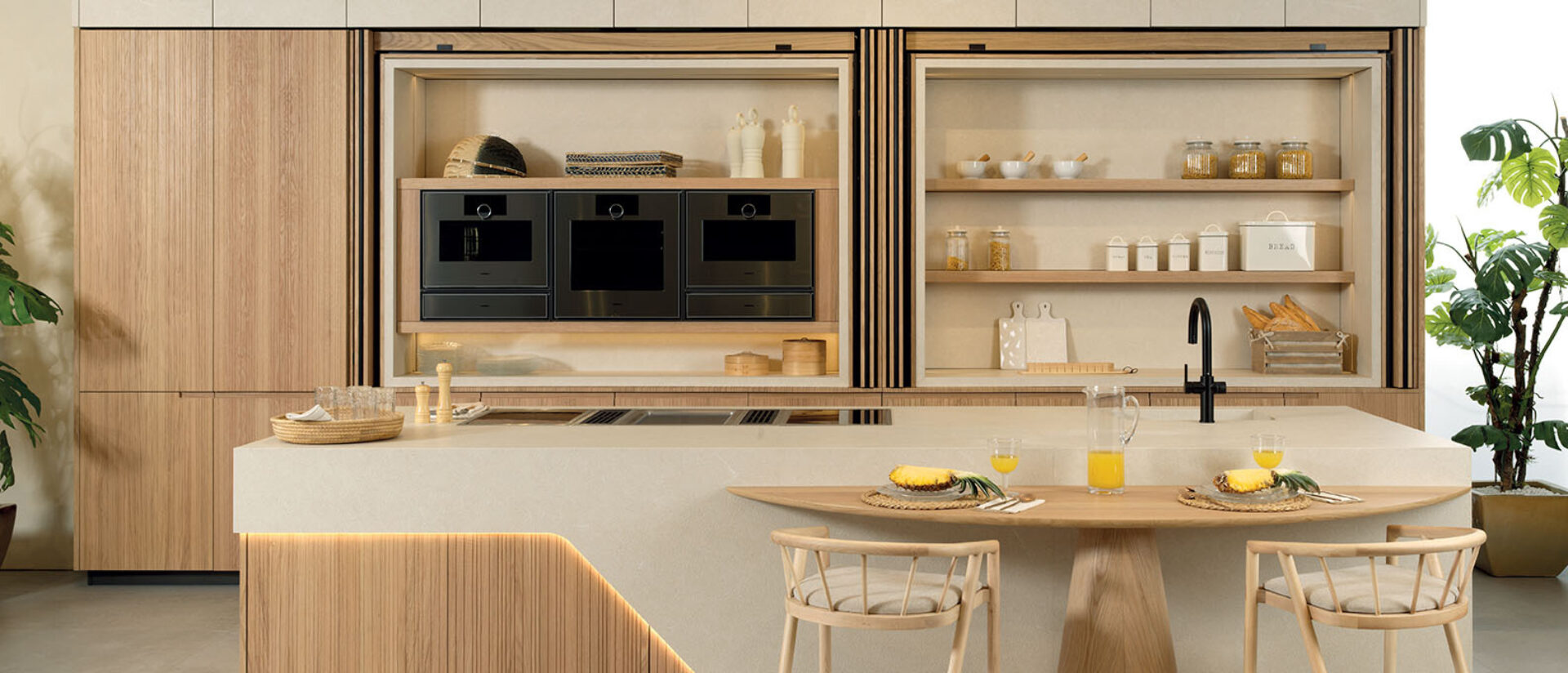 Modern minimalist kitchen with light wood cabinets and black ovens, open shelves, island bar with two stools, pineapple and orange juice.