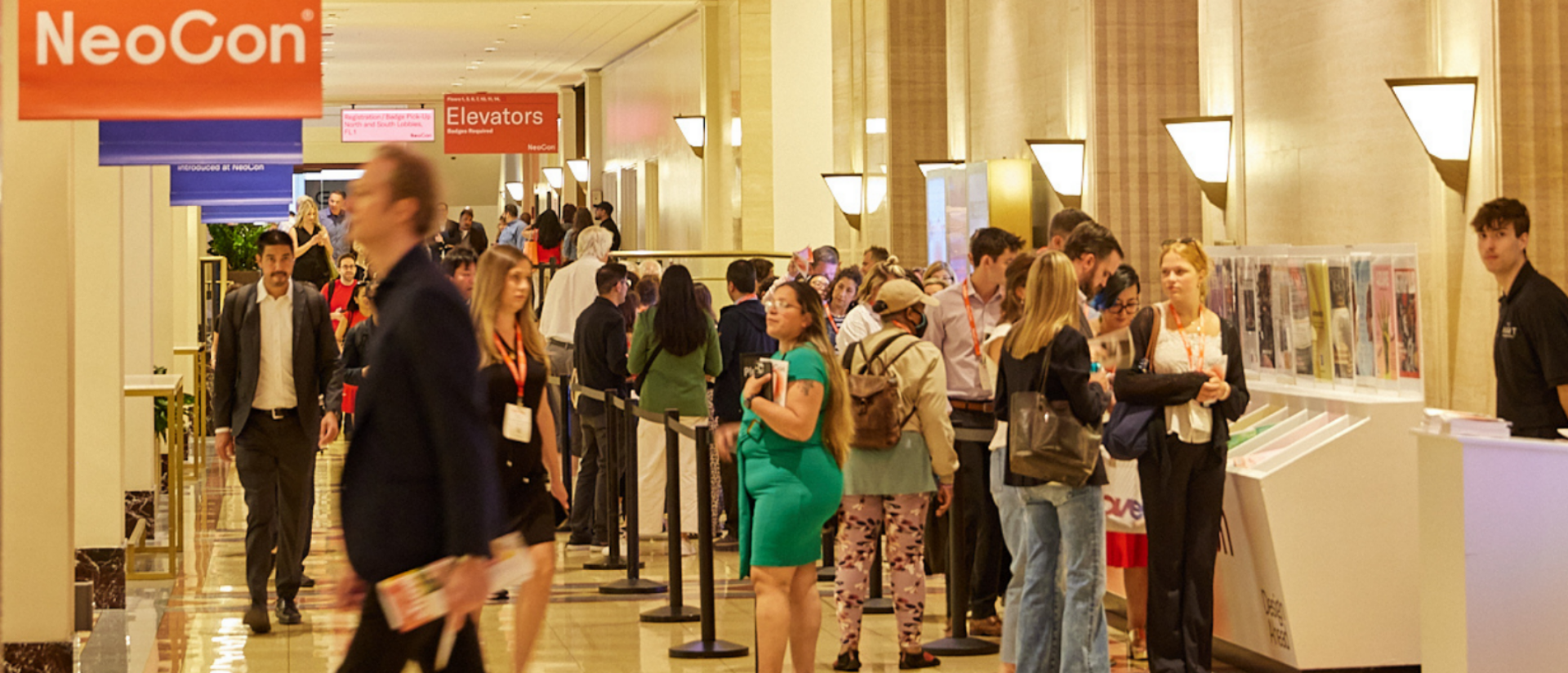 A bustling conference hall with attendees lined up near booths, signs indicating "Elevators" and "NeoCon," and informational displays nearby.