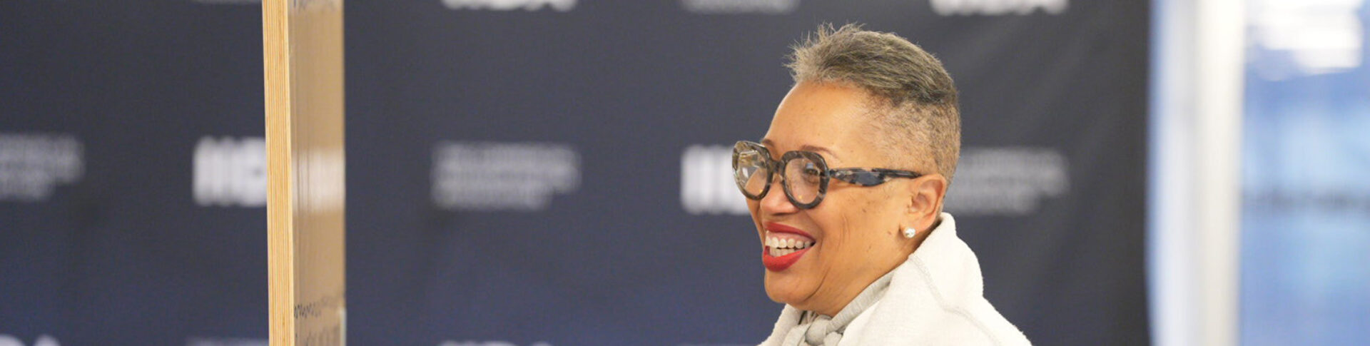 A smiling woman with short grey hair and large glasses stands next to a wooden board, set against a dark background with text.