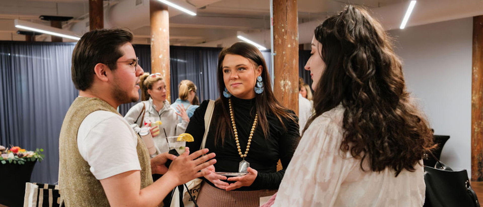 A group of three young adults engages in conversation at a trendy event space, with drinks in hand and a lively background.