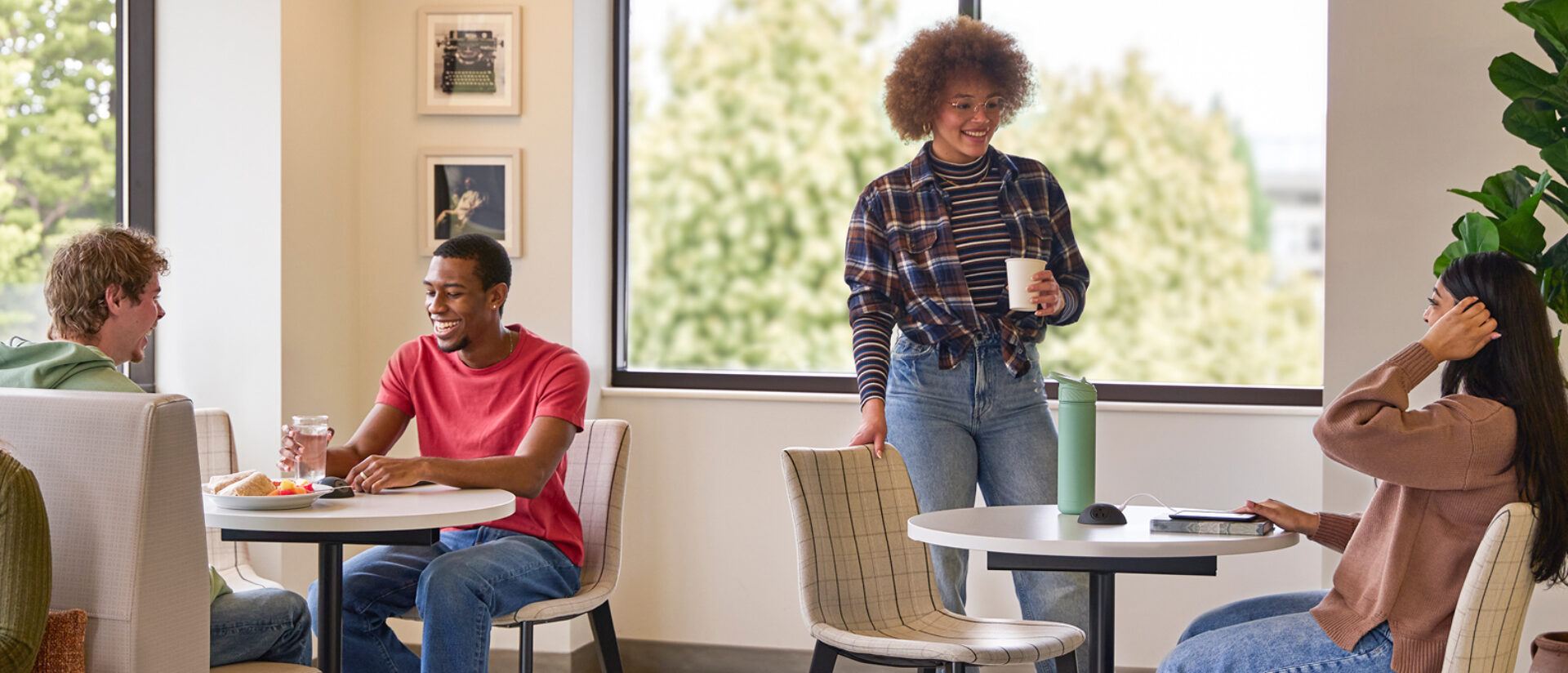 Four young adults in a sunny lounge: man in a red tee laughs with a friend at a table, woman in plaid holds coffee, another tucks hair.
