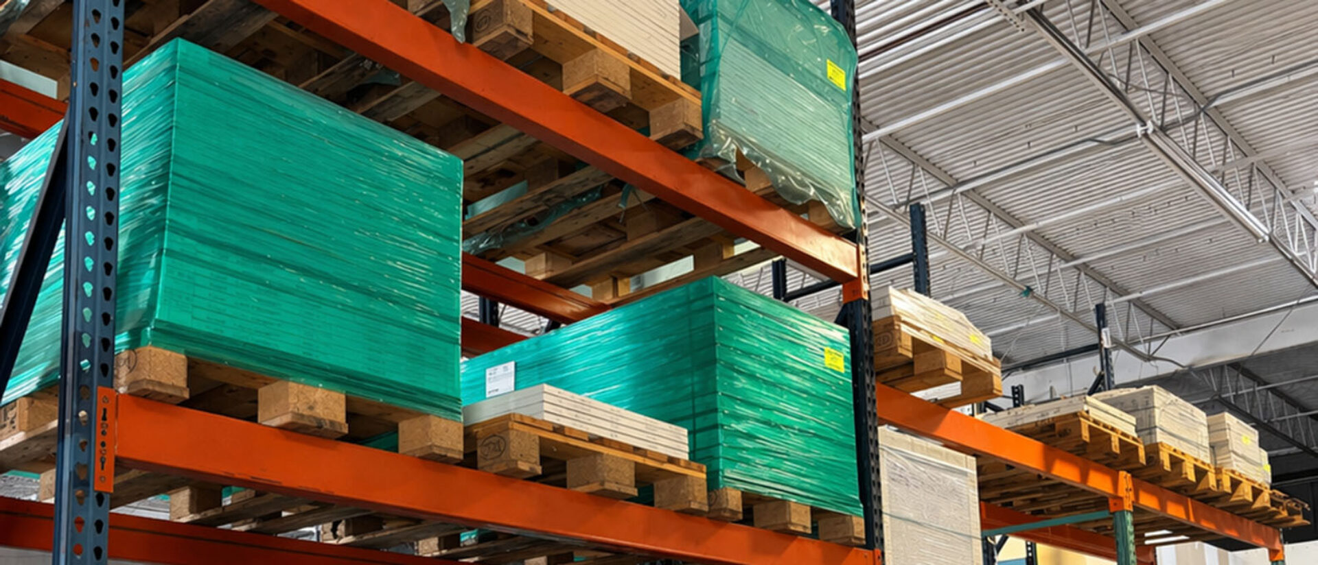 Stacks of wooden pallets display sheets of green and beige materials in a spacious warehouse with high ceilings and industrial shelving.