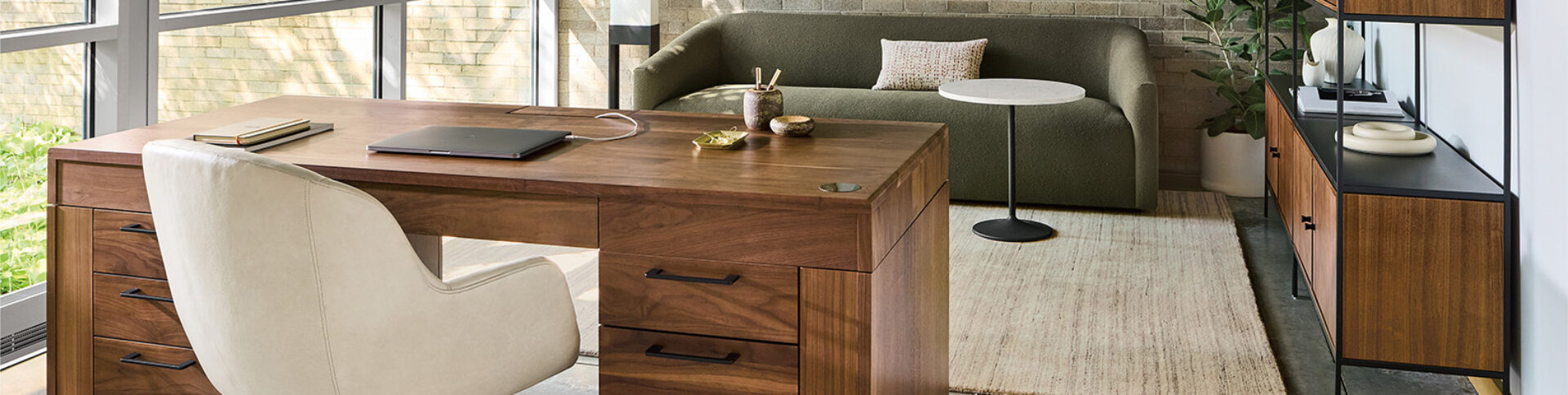 Sunlit modern home office with a walnut desk and laptop, cream leather chair, olive sofa, round marble side table and wood shelving.