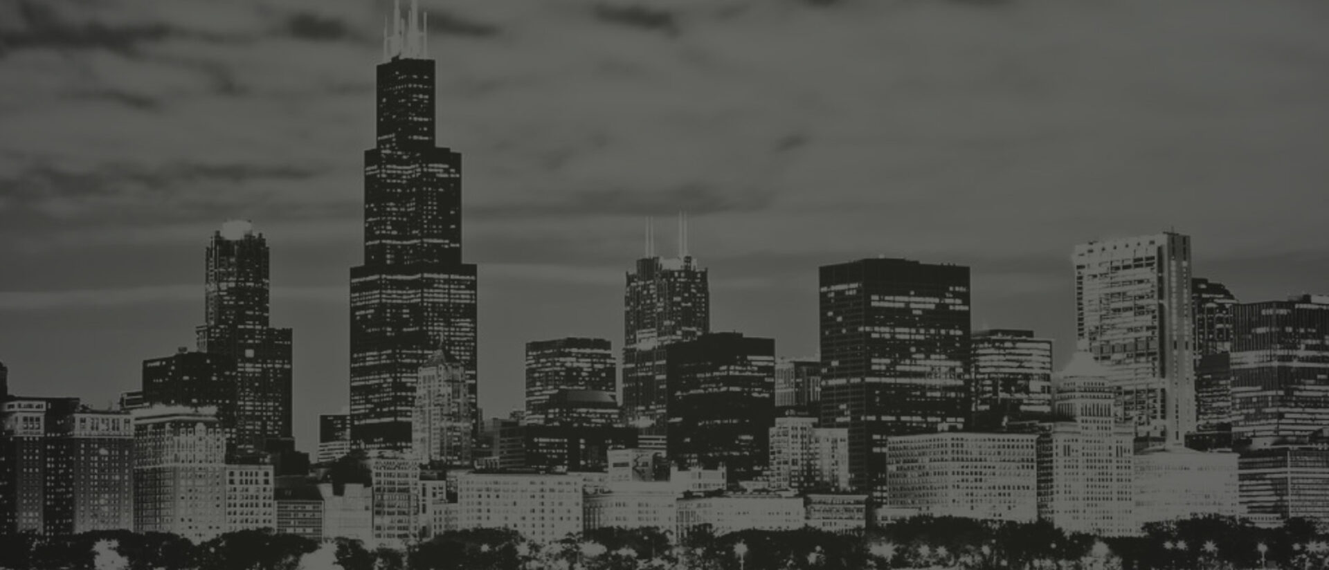 Black-and-white nighttime skyline of Chicago with the Willis Tower rising above illuminated downtown skyscrapers, cloudy sky and lit trees.