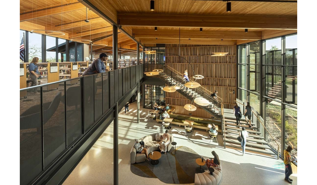 Sunlit two-story library with warm wood ceiling, mezzanine balcony, floating central staircase, round pendant lights and seating clusters.