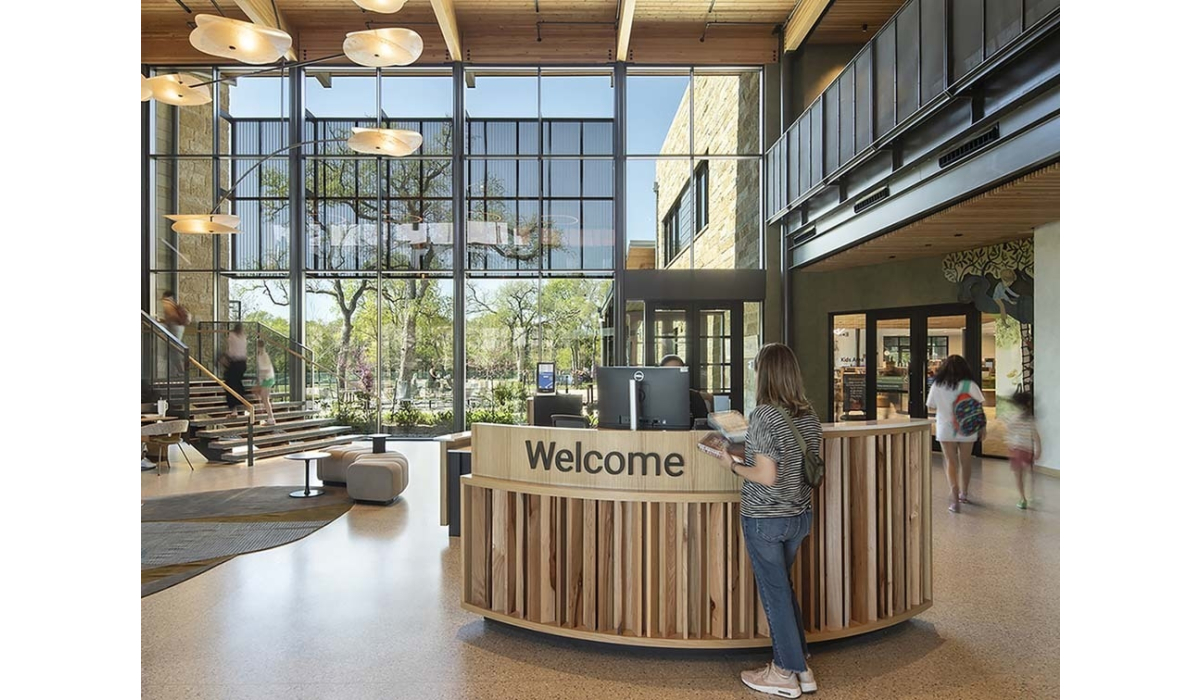 Sunlit modern library lobby with tall glass wall, wooden curved welcome desk, staircase to mezzanine, visitors walking and a woman at desk.