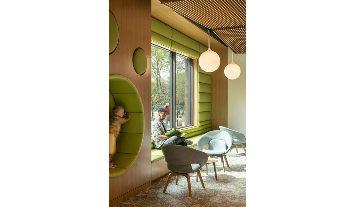 A sunlit reading nook with green window seat and circular alcove where a child climbs while a man reads, pendant lights overhead.