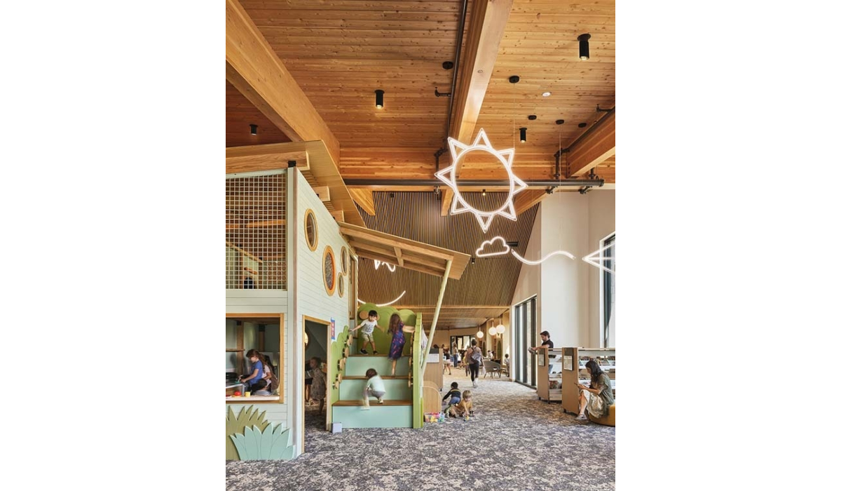 Bright wooden community playroom with high beamed ceiling, neon sun decor, green play stairs where children climb and adults supervise.