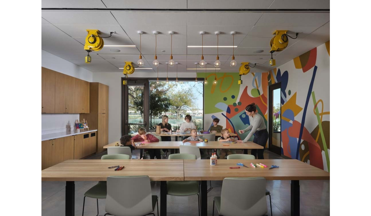 Bright art classroom with children and two adults at long tables, colorful abstract mural on wall, pendant bulbs and yellow ceiling reels.