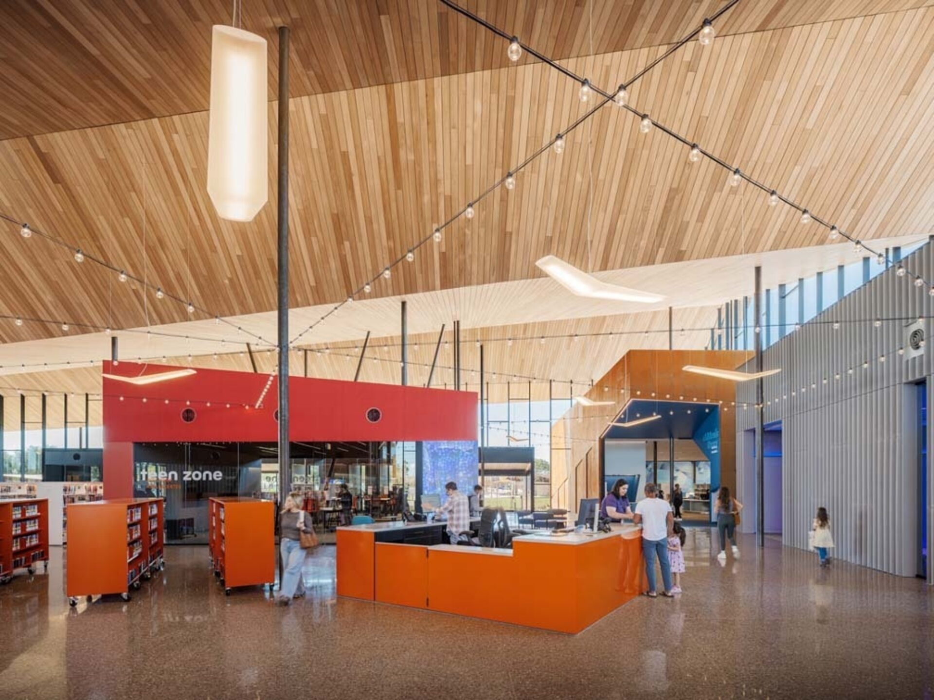 Sunlit modern library interior with a slatted wood ceiling, string lights, orange service desk, red teen-zone pod, book carts and visitors.