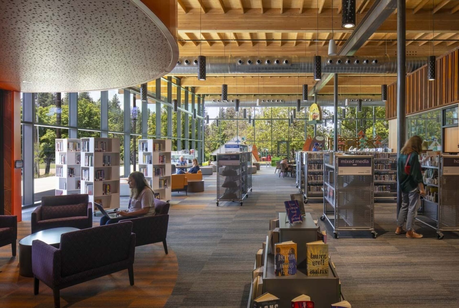 Sunlit modern library with tall glass windows, exposed wooden beams, mobile book carts, lounge seating and patrons browsing amid trees.