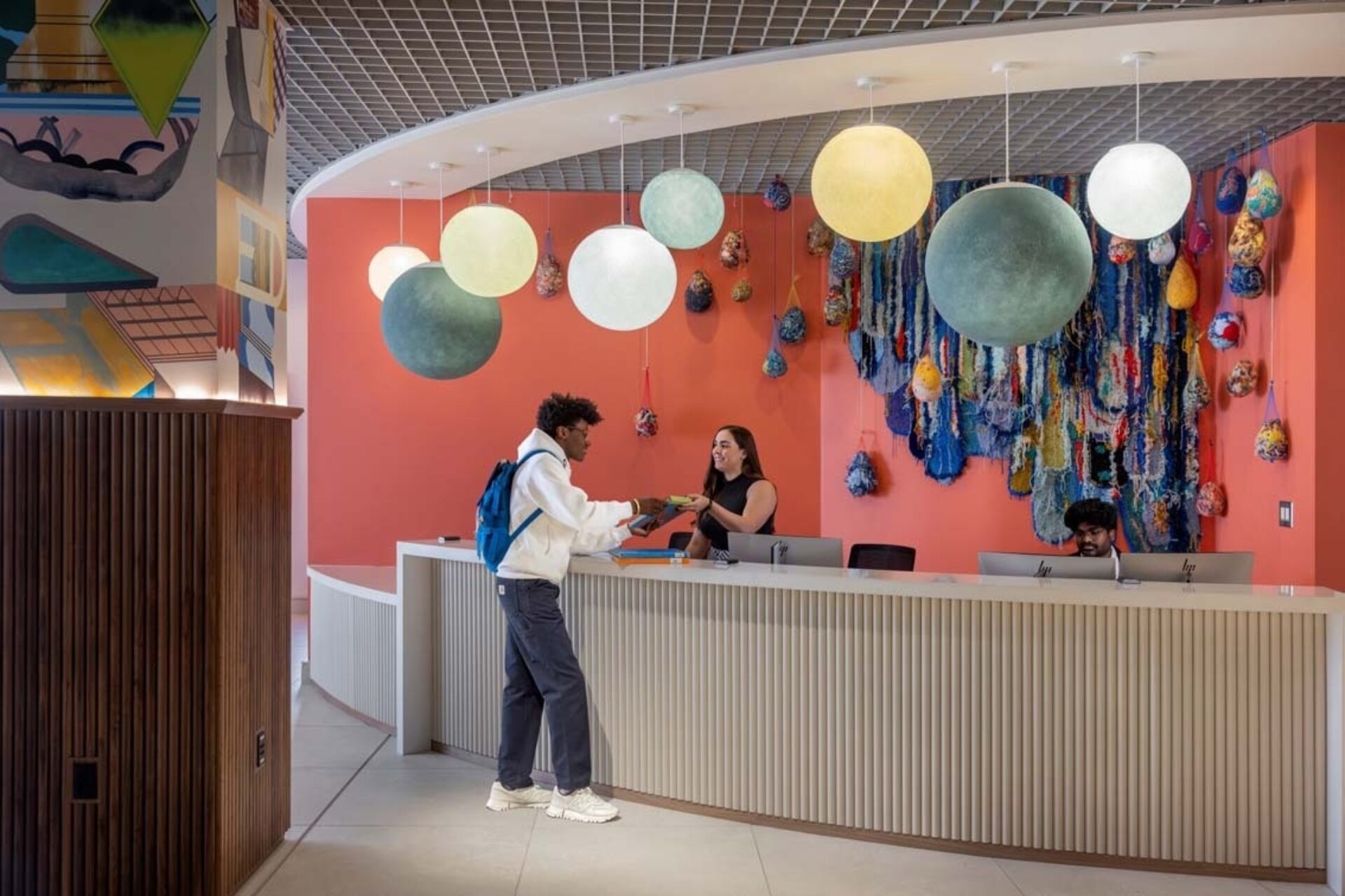 Student with blue backpack hands documents to a smiling staff at a curved desk under globe pendant lights and colorful textile wall art.