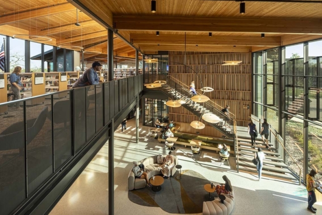 Sunlit two-story library with warm wood ceiling, mezzanine balcony, floating central staircase, round pendant lights and seating clusters.