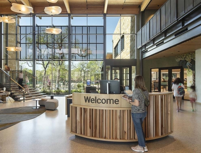 Sunlit modern library lobby with tall glass wall, wooden curved welcome desk, staircase to mezzanine, visitors walking and a woman at desk.