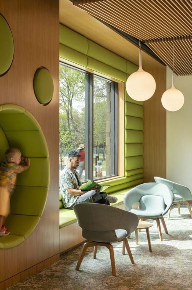 A sunlit reading nook with green window seat and circular alcove where a child climbs while a man reads, pendant lights overhead.