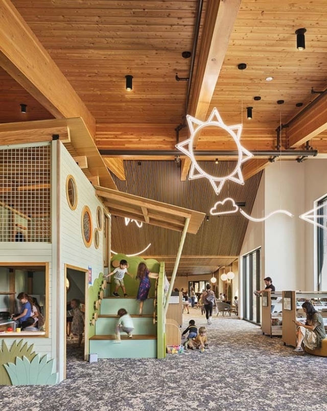 Bright wooden community playroom with high beamed ceiling, neon sun decor, green play stairs where children climb and adults supervise.