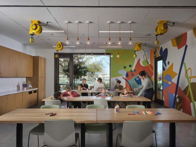 Bright art classroom with children and two adults at long tables, colorful abstract mural on wall, pendant bulbs and yellow ceiling reels.
