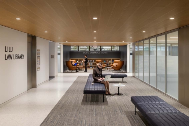 Spacious modern law library lounge with leather armchairs, shelving, low tufted benches and a woman in business attire typing on a laptop.