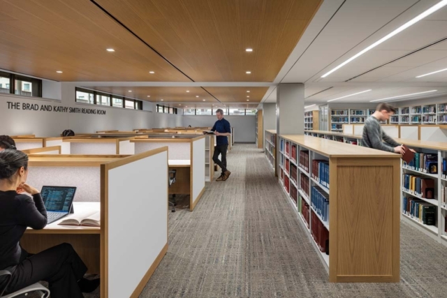 Spacious modern reading room with wood-trimmed study carrels on the left, tall bookshelves on the right, and students studying and browsing.