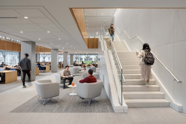Bright modern library interior with wide staircase on right, people studying and chatting in lounge seating and arched windows beyond.