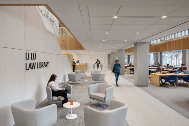 Spacious modern LIU Law Library lobby with marble wall sign, gray lounge chairs, a student on a phone, people studying at wooden tables.