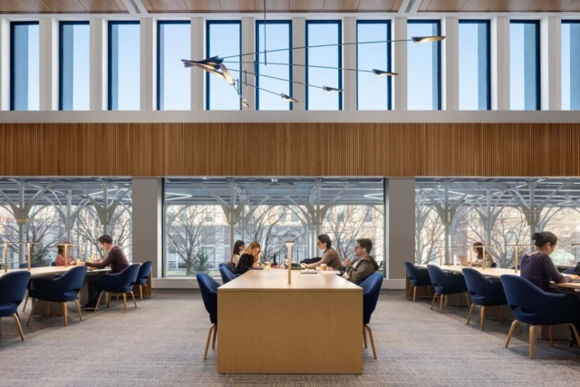 Spacious library room with a long wooden study table, blue chairs, people studying beneath tall windows and sculptural hanging lights.