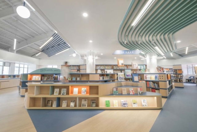 Bright modern library interior with tiered wooden display shelves, teal curved ceiling slats, rows of books and a blurred person walking.