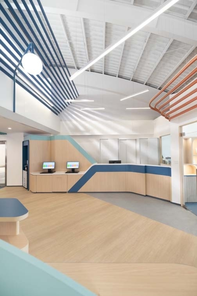 Bright reception area with curved light wood desk and blue trim, two computer kiosks, vaulted white ceiling and linear pendant lights.