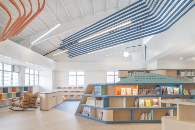 Bright modern kids' library with curved blue ceiling slats, tiered orange-and-blue seating, rounded wooden bookshelves and leather rocker.