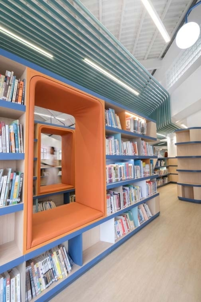 Modern library interior with wood floors, long blue wood shelving filled with books, inset orange reading nooks and teal ceiling slats.