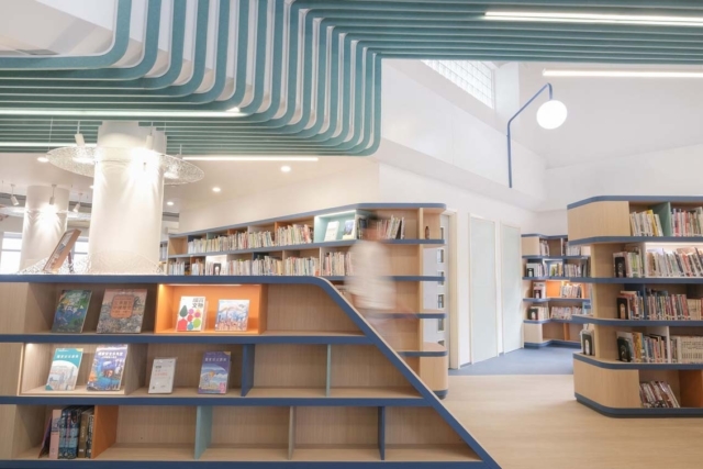 Bright modern library with curved teal ceiling slats, rounded wooden shelves with blue trim, display books and a blurred person walking.