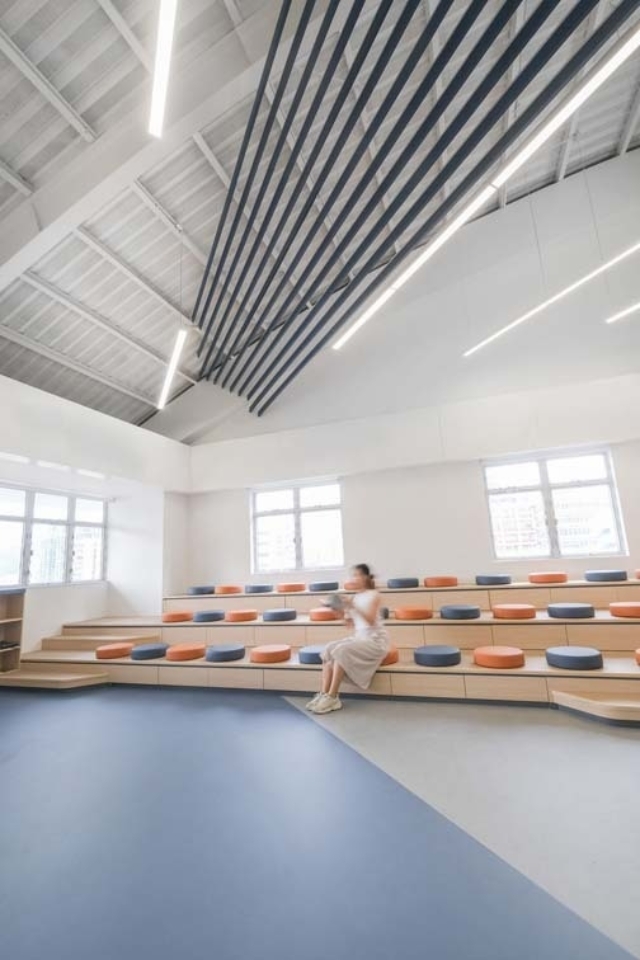 Bright room with tiered wooden seating topped by orange and gray cushions, ceiling with black slats and lights, blurred woman seated.