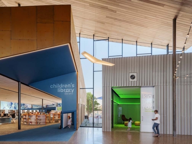 Bright children's library entrance with a blue angular canopy, wooden ceiling, glass walls, green playroom and two people entering.