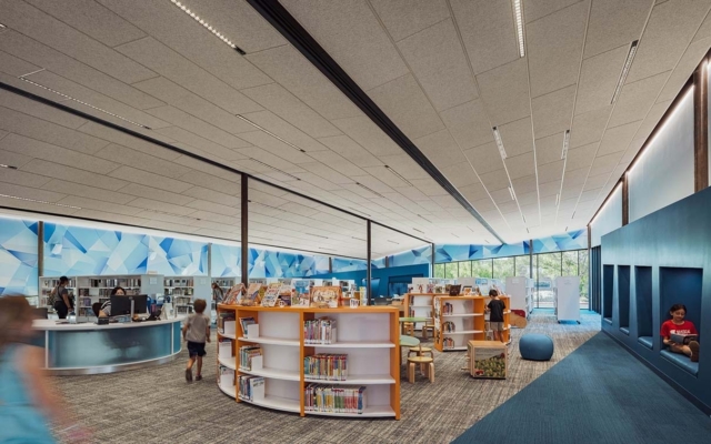 Bright children's library area with curved low white and orange shelves, kids browsing, blue geometric mural, window cubbies and seating.