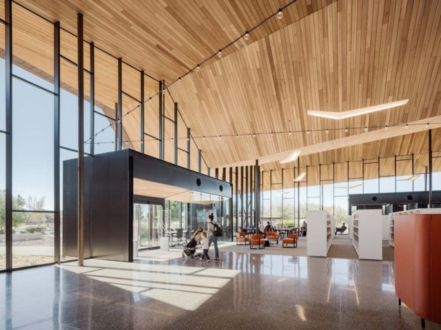 Spacious modern library with vaulted wood ceiling, tall windows, polished floors, shelves and orange lounge chairs, someone with a stroller.