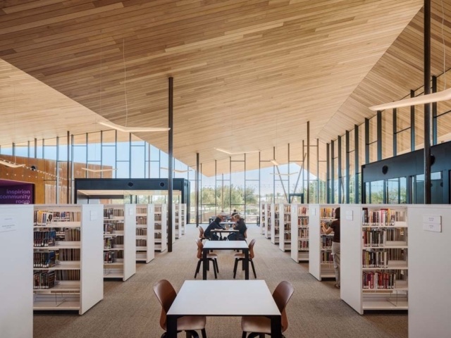 Spacious modern library with a high wood-paneled vaulted ceiling, floor-to-ceiling windows, rows of white bookshelves and patrons studying at tables.