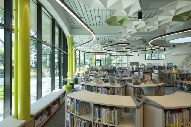 Bright children's library with low curved shelves, tall windows, lime-green columns, geometric ceiling panels and winding LED lights.