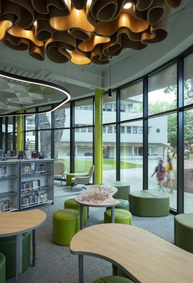 Modern school library with undulating wooden ceiling fixture, curved light ring, tall windows, green stools and sculpted curved tables.