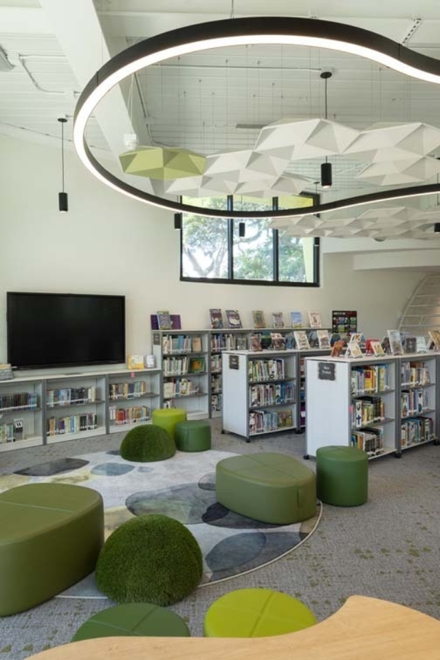 Bright modern children's library reading area with circular LED ring light, geometric ceiling panels, low bookshelves, and green round seats.