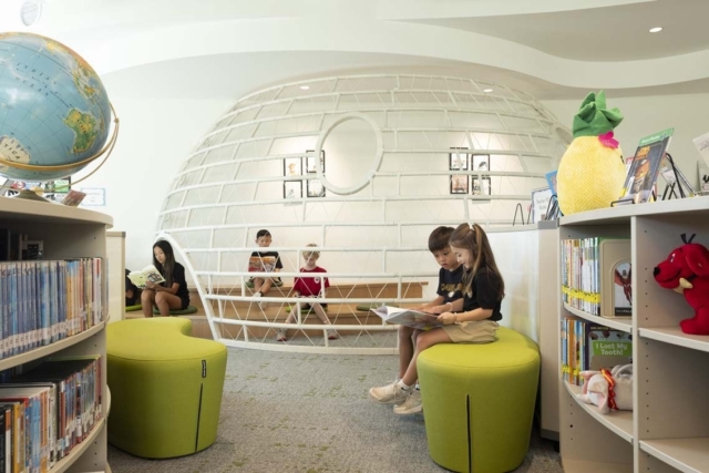Children read in a bright children's library featuring a white spherical wire reading pod, green round stools, globe and colorful shelves.