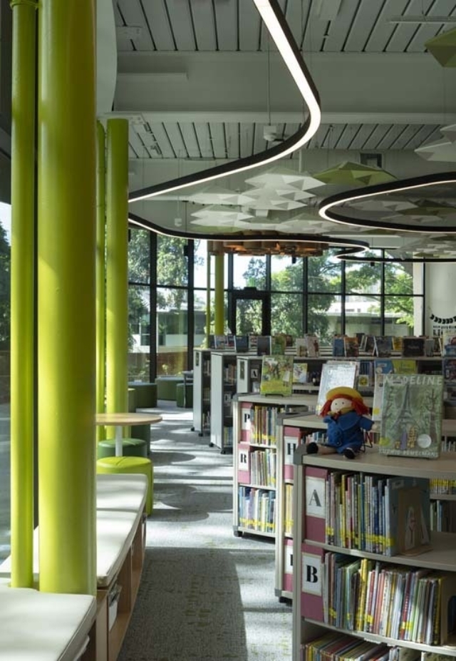 Bright kids' library section with lime green columns, curved LED lights, low shelves topped by a rag doll and tall windows overlooking trees.