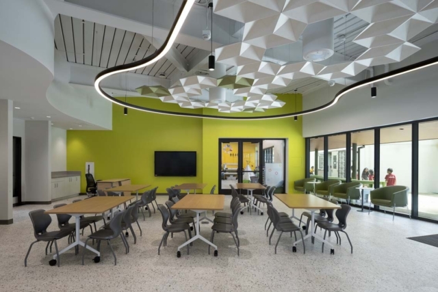 Modern learning space with lime green wall, geometric ceiling panels, circular LED ring, movable wood tables and gray chairs on casters.