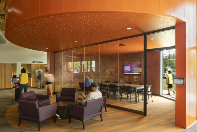 Modern library interior with an orange curved ceiling, glass-walled study room, four purple armchairs in a circle and patrons using devices.