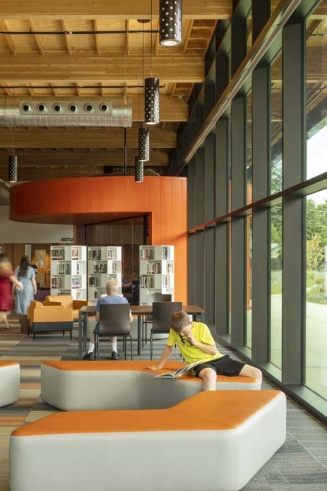 Sunlit library interior with floor-to-ceiling windows, orange curved wall and seating; boy in yellow reads on modular bench by the glass.