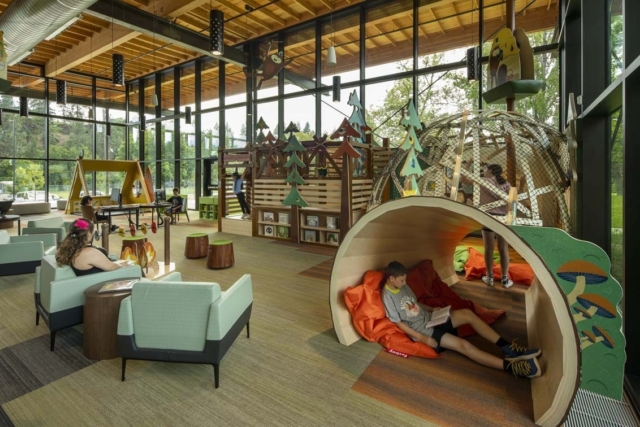 Bright glass-walled children's library with wooden ceiling, treehouse play structures, cozy tunnel nook where a boy reads on orange beanbags.