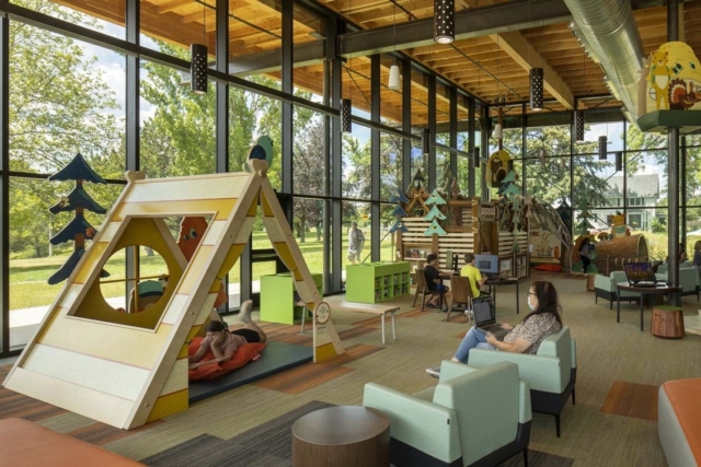 Bright children's library with floor-to-ceiling windows, wooden play structures and seating where kids play and adults work on laptops.